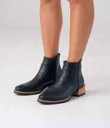 Bota corta de cuero vacuno color negro, con diseño de punta fina y cierre lateral. Presenta un taco bajo de madera y una entresuela de goma en tono natural que contrasta con el cuerpo de la bota.