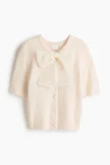 Saco de punto suave color crema, de manga corta y cuello redondo. Presenta cierre frontal con botones forrados y una moña decorativa de organza transparente en el escote. Posee terminaciones acanaladas en puños y ruedo.