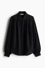 Blusa negra oversized en tejido liviano, con cuello clásico, tapeta con botones ocultos, canesú doble con frunces en el delantero y la espalda, y mangas globo largas con puños abotonados.
