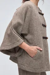 Capa de lana fina sin teñir, con diseño de corte amplio y mangas tipo kimono. Presenta cierre frontal mediante tres alamares de cuero en tono marrón.