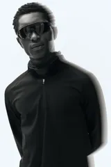 Remera deportiva negra de manga larga con cuello elevado y medio cierre frontal. Presenta mangas raglán superlargas con abertura para el pulgar.