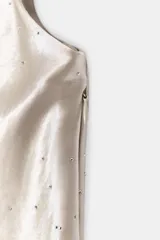 Top satinado color beige con cuello fluido, manga sisa y aplicaciones de brillos. Presenta falsos botones forrados en el lateral, bajo asimétrico con abertura y cierre de cremallera oculta.