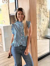 Camisa sin mangas de diseño a rayas celestes, marrones y blancas, con cuello alto fruncido, cierre frontal con botones y detalles de volados a lo largo de la botonera, en las sisas y en el ruedo.
