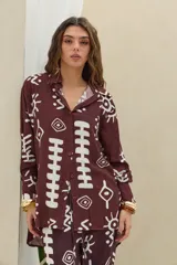 Camisa de manga larga color marrón oscuro con estampado gráfico en color blanco de símbolos abstractos, ojos y soles. Posee cuello camisero y cierre frontal con botones.