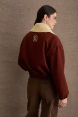 Campera tipo bomber de paño color marrón, con cierre frontal, puños y ruedo elásticos. Presenta un cuello de piel sintética desmontable en color crema y el logo de la marca bordado en el pecho.