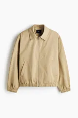 Campera tipo bomber de sarga de algodón en color beige, con cuello inglés, cierre frontal metálico y bolsillos diagonales ribeteados. Presenta terminaciones con elástico en los puños y el ruedo.