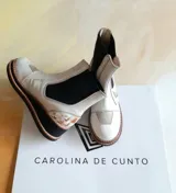 Botas cortas Carolina De Cunto modelo Kiria, confeccionadas en cuero de color blanco con detalles pintados a mano. Poseen inserciones elásticas laterales en color negro, suela de goma con plataforma y detalles texturizados en el talón.