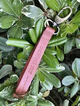 Llavero de cuero en formato de tira con diseño minimalista, confeccionado en cuero genuino marrón con interior en contraste color fucsia, pespuntes rosados y herrajes metálicos envejecidos con gancho tipo mosquetón.