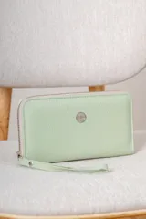 Billetera tipo sobre color verde menta con textura granulada, cierre perimetral y correa de muñeca extraíble. Presenta un aplique metálico circular con logo grabado en el centro.