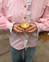 Camisa de manga larga abullonada en color rosa con rayas verticales finas. Presenta un diseño con volados en el cuello y a lo largo de la botonadura frontal, puños con detalle de rayas contrastantes y botones decorativos dorados.