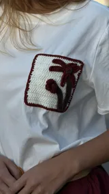 Remera blanca de corte boxy, con cuello redondo y manga corta. Presenta un bolsillo en el pecho con detalle de crochet con forma de palmera.