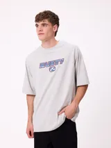 Remera de corte holgado color gris melange, con mangas raglán y estampado frontal con el logo de la marca Rusty en tonos azul y rojo.