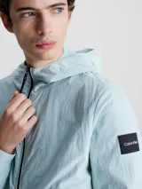 Campera con capucha Calvin Klein, confeccionada en nailon 100% reciclado color celeste. Tiene cierre de cremallera frontal, cuello alto, bolsillos delanteros con cremallera, puños y dobladillo elásticos, tirador de cremallera con la marca e insignia con el logo de Calvin Klein en la manga.