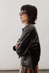 Chaqueta de cuero negra con cuello redondo, cierre frontal de botones forrados y bolsillos ribeteados con detalles de costura decorativa.