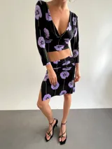 Falda midi de lycra con estampado floral en tonos violeta sobre fondo negro. Presenta un diseño con caderín fruncido y un tajo lateral en el ruedo.