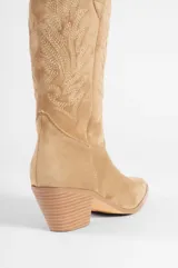 Bota de caña media color beige, confeccionada en suede, con pespuntes decorativos en la caña y la puntera. Presenta un tacón de bloque y punta en pico.