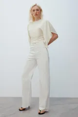 Camiseta tipo capa oversize de color blanco, confeccionada en hilatura de algodón 100%. Presenta cuello redondo, manga corta y un diseño de bajo asimétrico con caída pronunciada en uno de los laterales.