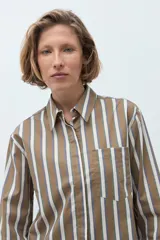 Camisa de corte clásico con estampado de rayas verticales en tonos khaki, blanco y azul. Presenta cuello camisero, cierre frontal con botones, bolsillo en el pecho y mangas largas con puños abotonados.