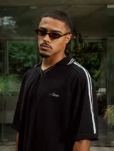 Chomba de tejido de punto cerrado color blanco, con cuello polo y mangas cortas. Presenta un bordado sutil en el frente y tres franjas negras en los hombros.