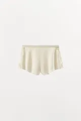 Shorts de tiro alto confeccionado en tejido satinado color crema, con cintura elástica, bajo con volantes y aberturas laterales decoradas con aplicaciones de encaje a tono.