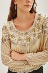 Camisa de manga larga en color crema, con diseño bordado en hilos dorados y plateados que forman líneas verticales y detalles florales en el pecho. Presenta un estilo elegante y sofisticado con aplicaciones brillantes.