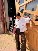 Camisa blanca con volumen, de manga larga, con bolsillo frontal grande decorado con un bordado rojo de un caballo y cierre frontal de botones oscuros.