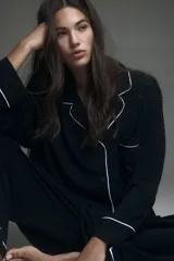 Pijama de dos piezas negro, compuesto por una camisa de manga larga con solapas de muesca, ribetes blancos y cierre de botones, y un pantalón de pierna ancha con elástico revestido en la cintura y ribetes blancos en los puños.