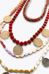 Pack de cinco collares superpuestos con cadenas de diferentes grosores. Incluye collares con cuentas rojas, marrones y amarillas, cuentas de cristal transparentes/rojas, y dijes metálicos circulares con textura martillada dorada y otros con diseño de espiral plateado/negro.