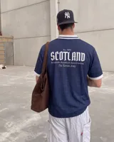 Camiseta de fútbol de Escocia de manga corta, color azul marino con diseño a cuadros sutiles, cuello en V y cuello camisero blanco con líneas azules. Presenta detalles estampados en el frente, incluyendo el escudo de la Asociación de Fútbol de Escocia, un león rampante y el texto 'Scottish Football Association'.