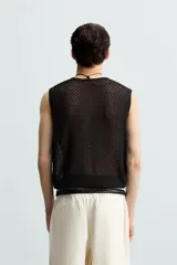 Chaleco de punto negro, con cuello redondo y manga sisa, confeccionado en tejido con estructura abierta (calado). Presenta bajo con aberturas laterales y acabados en rib.