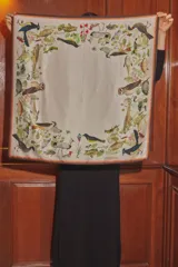 Pañuelo cuadrado de seda con estampado botánico y de fauna silvestre, que incluye ilustraciones de aves, peces y plantas sobre un fondo beige con bordes en tono café.