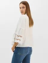 Camisa negra de poliéster con cuello camisero, escote en V y cierre frontal de botones. Cuenta con mangas anchas de corte acampanado y detalles de bordado suizo calado en las mangas y en el ruedo inferior.