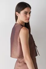 Top sin mangas de estilo cruzado, confeccionado en tela satinada color cobre. Presenta un diseño corto con cierre frontal mediante tiras finas para anudar y cuello redondo.