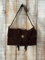 Cartera tipo bandolera de gamuza color marrón, con diseño rectangular, solapa frontal decorada con flecos colgantes y placa metálica distintiva. Cuenta con una correa larga para el hombro y costuras pespuntadas en contraste.