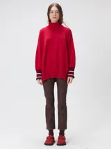 Pullover rojo de cuello alto, corte holgado, mangas largas y puños acanalados en tono burdeos con rayas blancas. Presenta un pequeño logo bordado en el pecho.