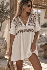 Vestido corto de lino color blanco, con escote en V y mangas cortas amplias. Presenta delicados bordados florales en tono marrón en la parte superior y en la línea del pecho, con terminaciones en festón.