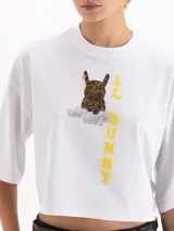 Remera blanca de corte cropped y manga corta, confeccionada en algodón. Presenta una estampa frontal con un conejo con estampado animal print sobre una nube y el texto 'IL BUNNY' en color amarillo.