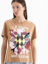 Remera de algodón color marrón con estampa frontal que incluye texto, flores, flechas y un patrón de rombos. Presenta cuello redondo y mangas cortas.