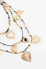 Collar de múltiples hebras negras con dijes colgantes dorados en forma de corazón de diferentes tamaños y texturas, intercalados con cuentas cilíndricas doradas.