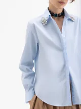 Camisa de algodón con lycra en color celeste claro, de corte clásico y manga larga. Presenta apliques de piedras de cristal en las puntas del cuello y una cartera oculta con botones.