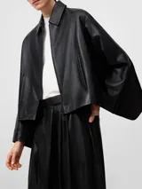 Chaqueta negra de piel ovina con diseño desestructurado, cuello solapa, cierre frontal con cremallera y bolsillos de vivo inclinados. Presenta una capelina en la espalda y mangas largas.