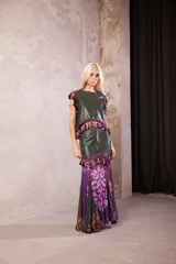 Falda larga de cuero verde oliva con diseño de paneles. Presenta una sección superior de cuero con un galón decorativo de borlas en tonos morados y verdes, y una sección inferior de tela fluida con estampado floral en tonos violeta, morado y verde.