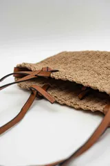 Bolso de mano tipo tote confeccionado en tejido texturizado efecto rafia en color beige, con asas dobles finas de cuero sintético marrón y detalles de nudos en los extremos.