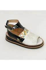 Sandalia de cuero Carolina De Cunto modelo Leyla, con semi plataforma y diseño combinado en charol negro, cuero blanco y una franja con detalles pintados a mano en tonos dorados y multicolores. Cuenta con tira ajustable en el talón con hebilla metálica.