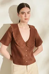 Blusa de lino color marrón con cuello redondo amplio tipo Peter Pan decorado con volados, mangas cortas y cierre frontal con botones.
