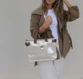 Bolso de mano tipo tote, confeccionado en cuero metalizado color plateado con detalles en cuero marrón en las asas y laterales. Cuenta con un bolsillo frontal con cierre giratorio y herrajes metálicos.