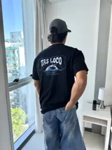 Remera negra de corte holgado con estampado gráfico en la espalda que dice "TAS LOCO" en letras rosas y celestes, con una ilustración de una ola y el texto "SUNNY WITH A CHANCE OF STOKE".