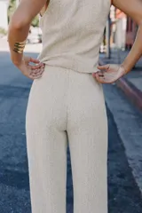 Pantalón de tiro medio, pierna ancha, confeccionado en tejido texturado de color claro con hilo metalizado. Presenta falsos bolsillos delanteros con botones metálicos dorados.