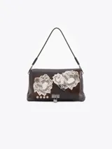 Cartera de hombro color marrón, confeccionada en cuero vacuno con detalles en descarne. Presenta un diseño con apliques de encaje, tachas metálicas y un cierre frontal tipo candado. Posee una estructura semirrígida y correa de cuero.