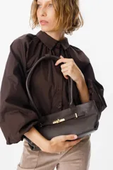 Bolso de hombro confeccionado en cuero vacuno color bordó, con textura granulada. Presenta un diseño estructurado con una tira decorativa frontal que incluye un cierre metálico tipo torniquete en tono dorado.